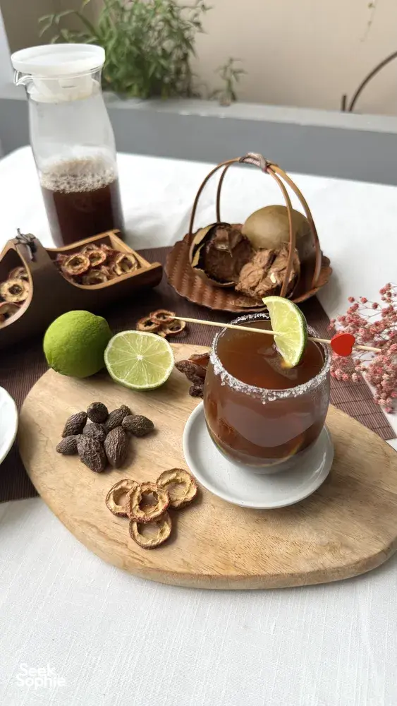 Mocktail gelap terinspirasi TCM dalam gelas dengan irisan jeruk nipis dan pengaduk merah, dengan herba kering, irisan akar, dan sirup di atas nampan di Singapura