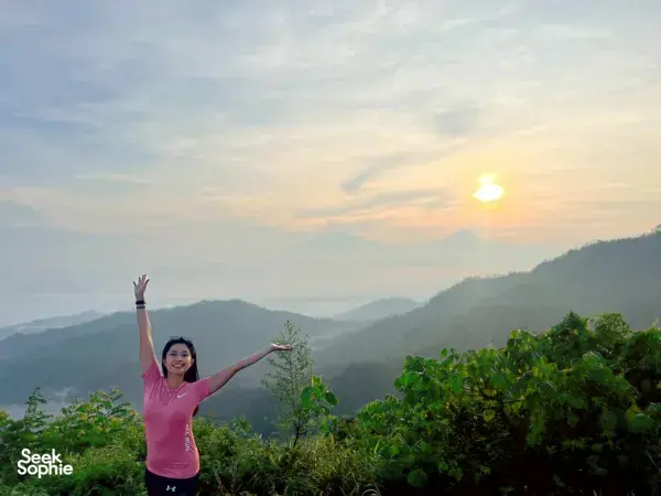 Woman watching sunrise over three volcanoes from a hillside viewpoint in Yogyakarta, with misty mountain ranges at dawn.