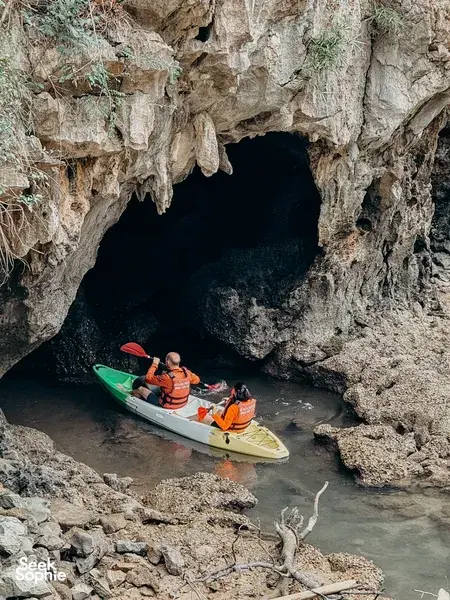 Kayakers in orange lifejackets drifting near a rocky cave entrance during a Hidden Lan Ha Bay stop, Cat Ba