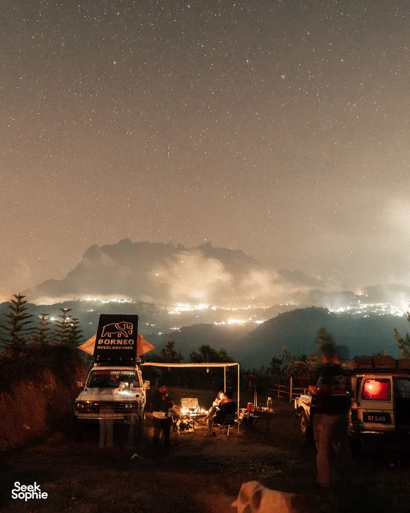 Starry night wild camping with a campfire and vans at a ridge, looking towards Mt Kinabalu from Kota Kinabalu