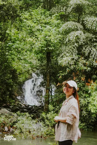 Woman in a white shirt and hat stands by a rainforest river, smiling as a waterfall spills into the pool in Khao Sok National Park