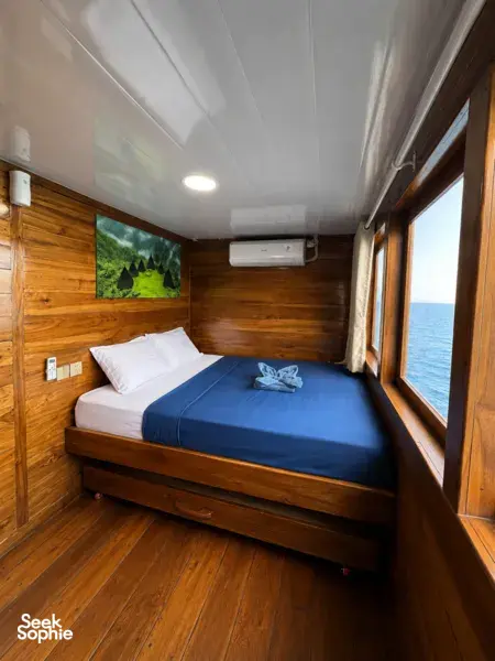 Wooden phinisi cabin with air-conditioned double bed, curtain-side window and wall art on a shared Komodo cruise from Labuan Bajo