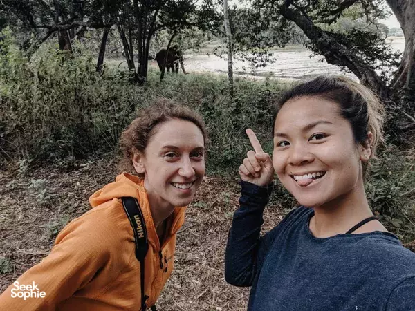Travellers smiling with peace-sign pose beside a safari vehicle as elephants walk in the trees near Anuradhapura