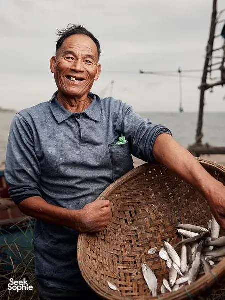 Ha smiling beside a woven basket of small fish at the coast in Hoi An, sharing local morning fishing traditions