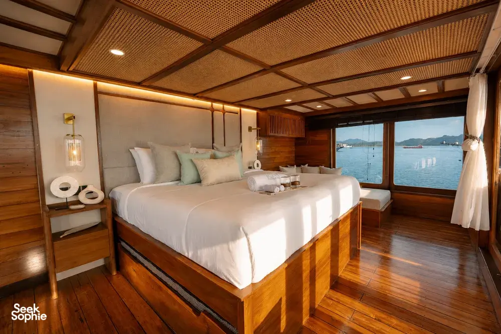 Wooden phinisi cabin with a made double bed, pillows, and seating by the window on a trip to Komodo Island