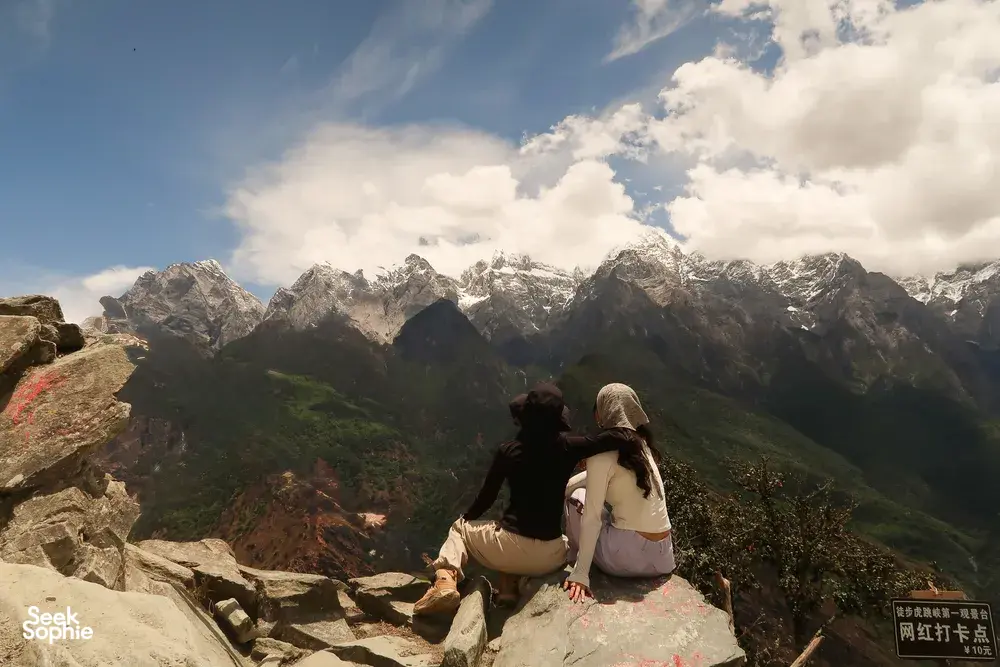 Two hikers sitting together on a viewpoint ledge, gazing at rugged snow mountains over Tiger Leaping Gorge, Lijiang, 2D1N high trail