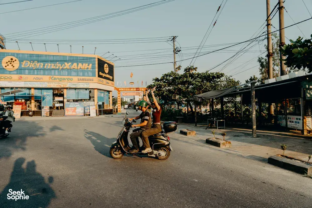 Dua pengendara Vespa mengangkat tangan saat melaju melewati deretan toko di Hoi An dalam perjalanan seru menyusuri jalan belakang bersama pemandu