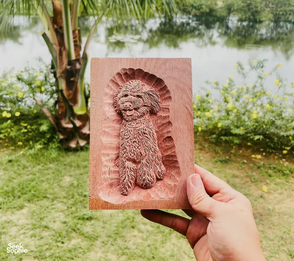 在会安户外,一只手拿着一尊雕刻木狮,背景是郁郁葱葱的花园景致