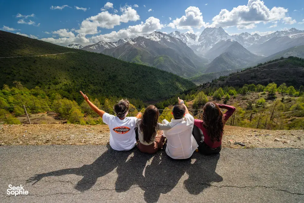 Travellers sitting on a roadside viewpoint above Shangri-La, arms raised toward snowy peaks during a 6-day Loop with a Tibetan guide