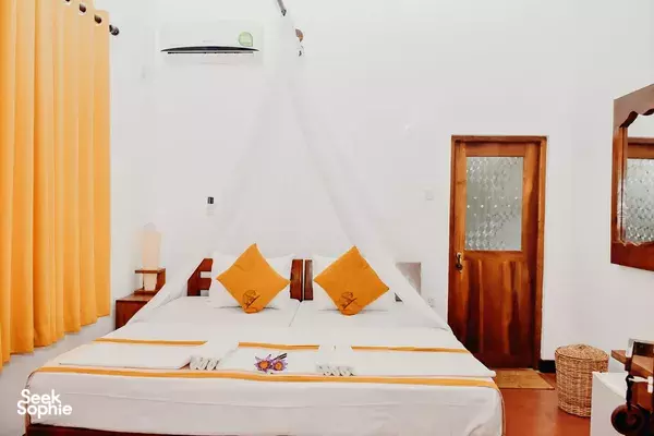 Sunlit cottage bedroom with two pillows and orange curtains at The Elephant Next Door in Udawalawe garden