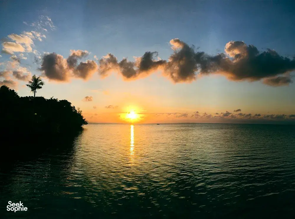 Sunset over calm ocean with a warm sun reflecting on the water and palm-lined coast seen from Semporna