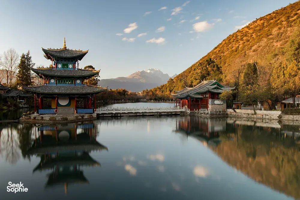 Traditional Chinese pagodas by a calm lake in China at sunrise, with mountains and golden hills reflected in the water