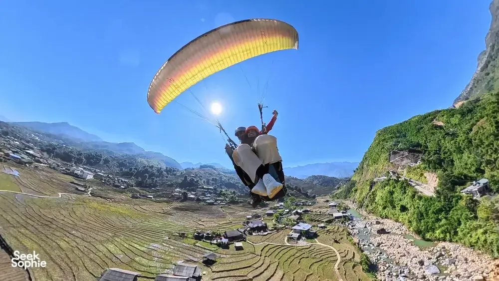 Paraglider gliding above layered terraced hills and a winding road through the valley, guided by Sapa Paragliding in Sapa, Vietnam
