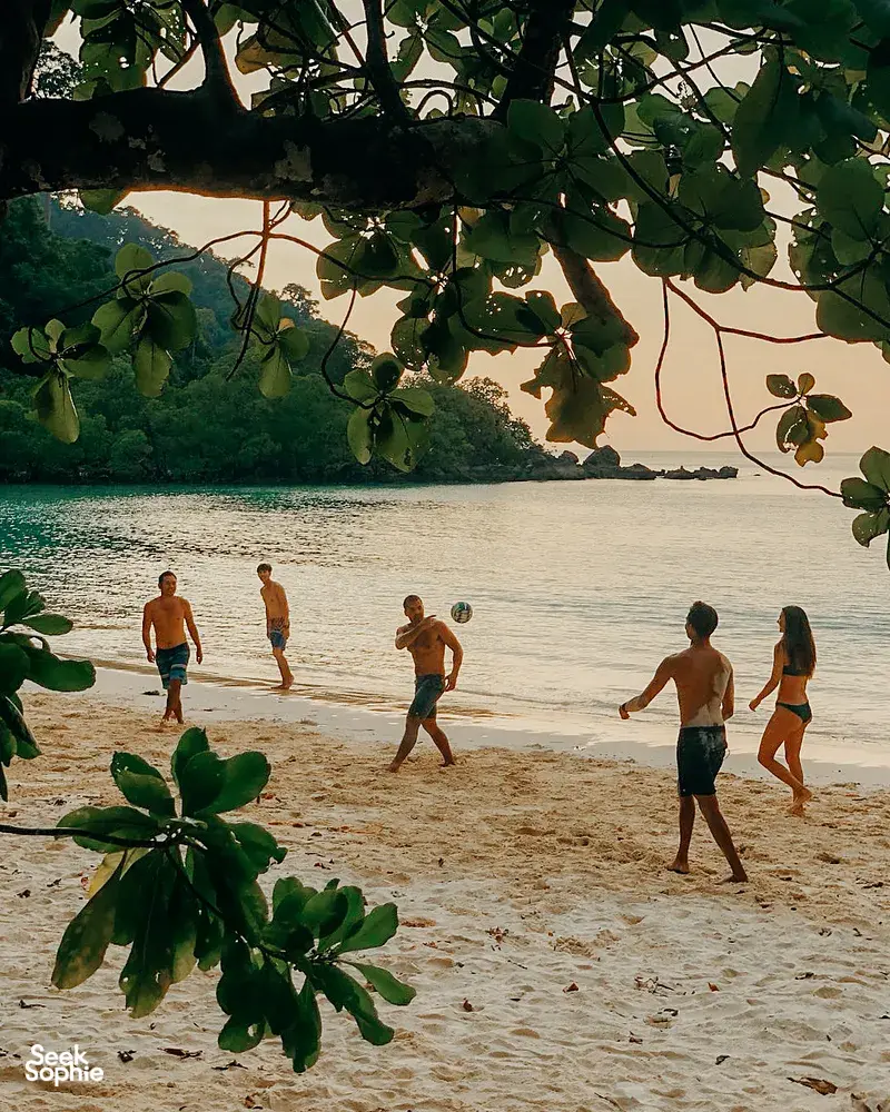People strolling on a sandy Surin Island beach at Khao Lak, framed by leafy branches, as they head toward the water
