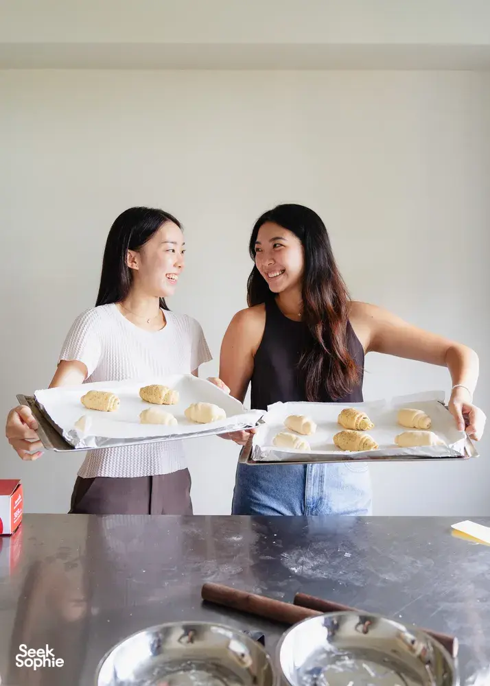 Two learners holding trays of shio pan buns while smiling at the counter, with rolled dough and bowls nearby during workshops in Singapore