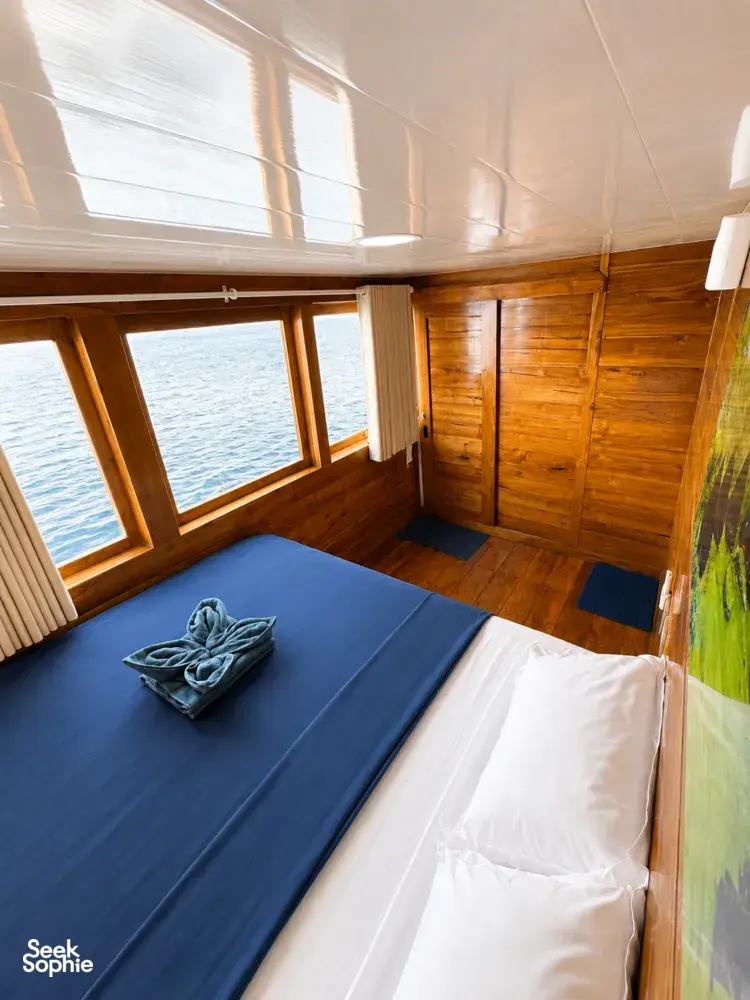 La Famille wooden phinisi cabin with two beds made up, large sea-facing windows and calm ocean outside on a Komodo cruise Labuan Bajo
