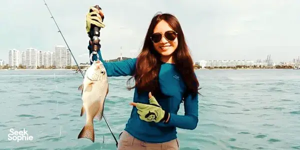 Woman on a Singapore fishing yacht holding up a fresh catch from the water, with the city skyline behind her