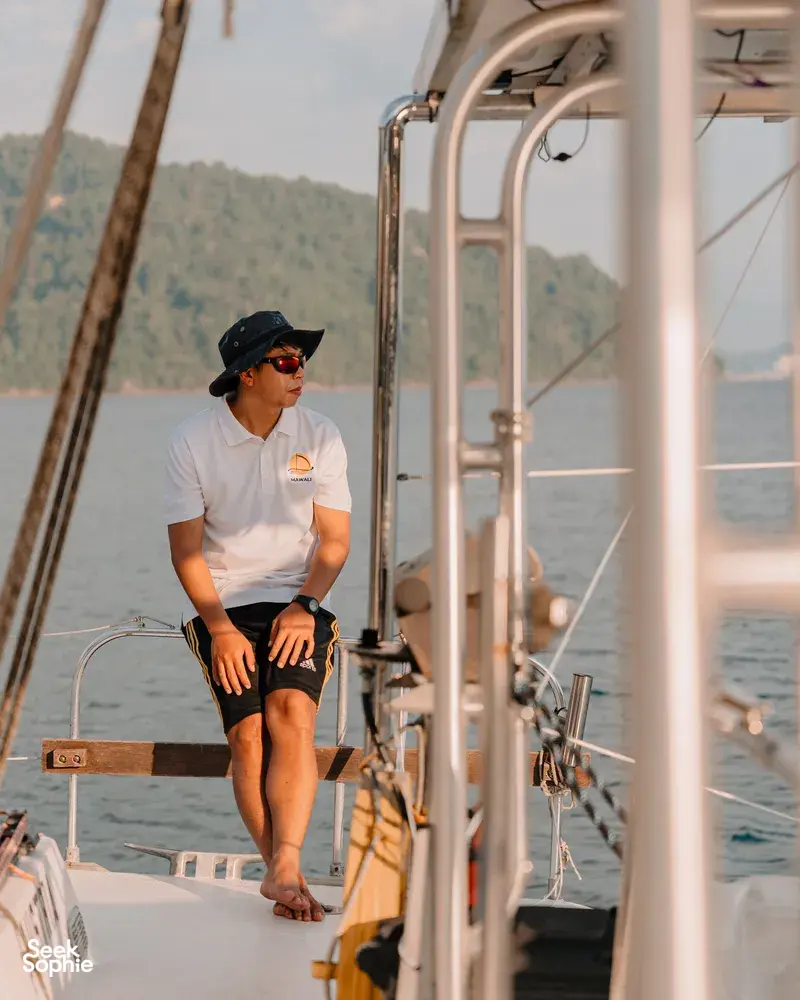 Crew member seated along the deck rail on SY Mawali catamaran as it sails past forested hills near Kota Kinabalu