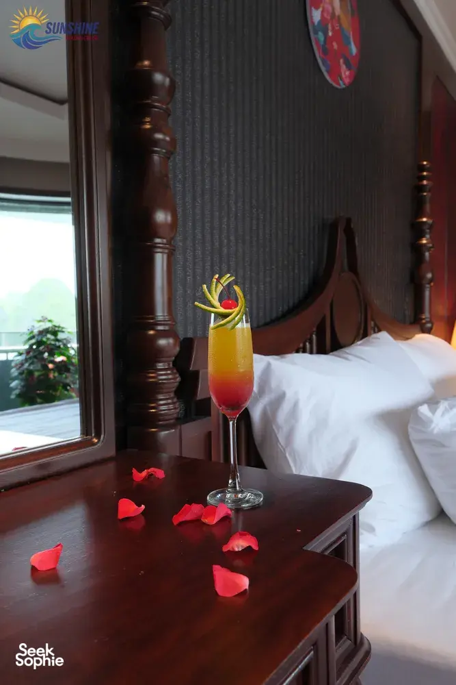 Cocktail with a flower garnish on a bedside table beside rose petals in a luxe Cat Ba cruise cabin by Sunshine Golden Cruises