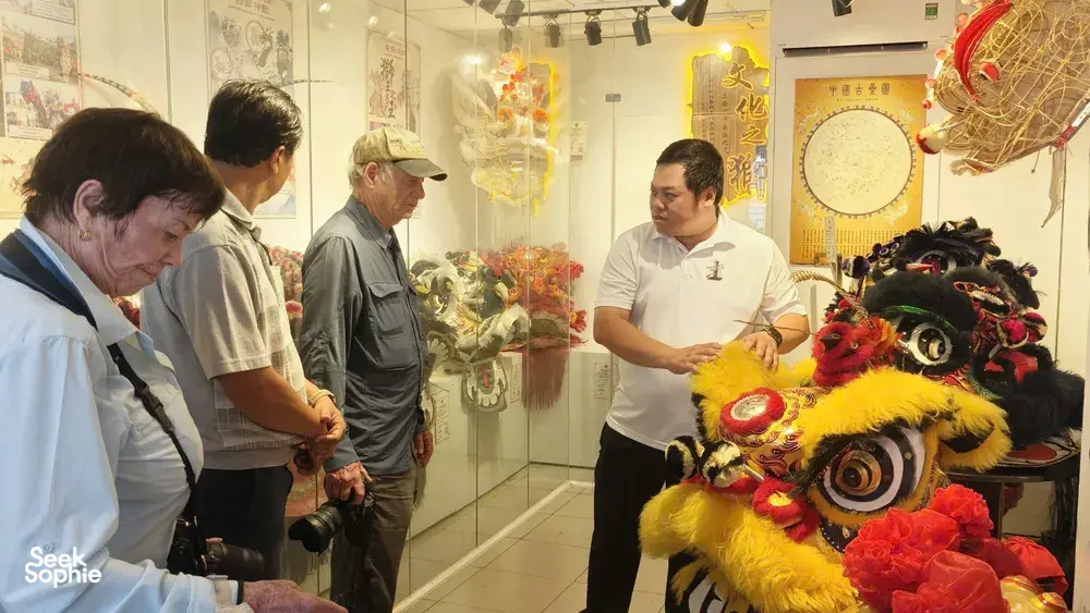 Museum visitors viewing a lion-dance mask display as a guide points at exhibits in Ho Chi Minh City Chinatown