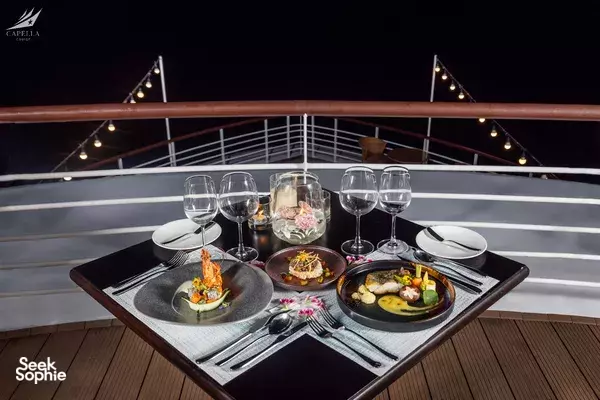 Evening dinner on a Halong Bay cruise deck with plated dishes, wine glasses and warm string lights above, Lyra Cruise Collection