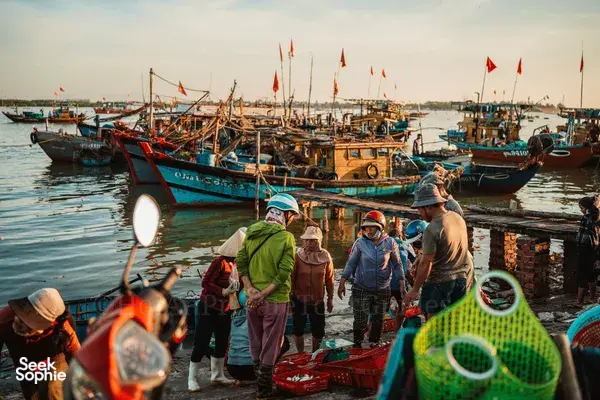 Small group of helmed travellers walking along a Hoi An harbour dock at sunrise, surrounded by fishing boats with flags overhead