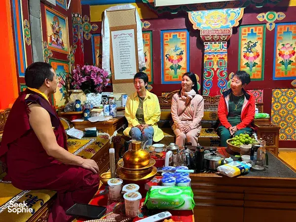 Lama in maroon robes leads a tea-time conversation as travellers sit by a decorated table of pots in Shangri-La