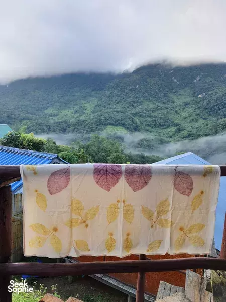 Leaf-pattern eco-print fabric laid out to dry on a wooden rail, with misty green Chiang Mai forest hills beyond