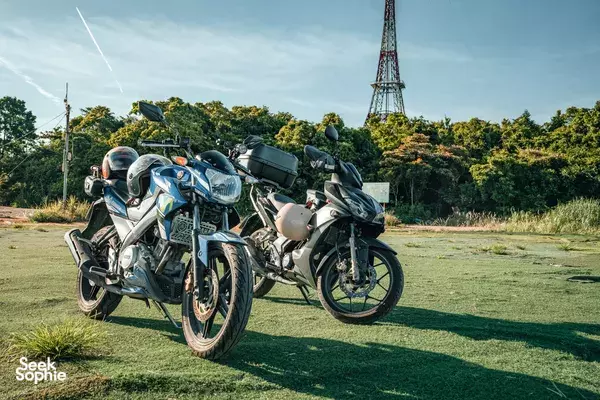 Parked dual motorbikes on a grassy roadside near a lattice tower in Da Nang, ready for a sunset motorbike wildlife adventure