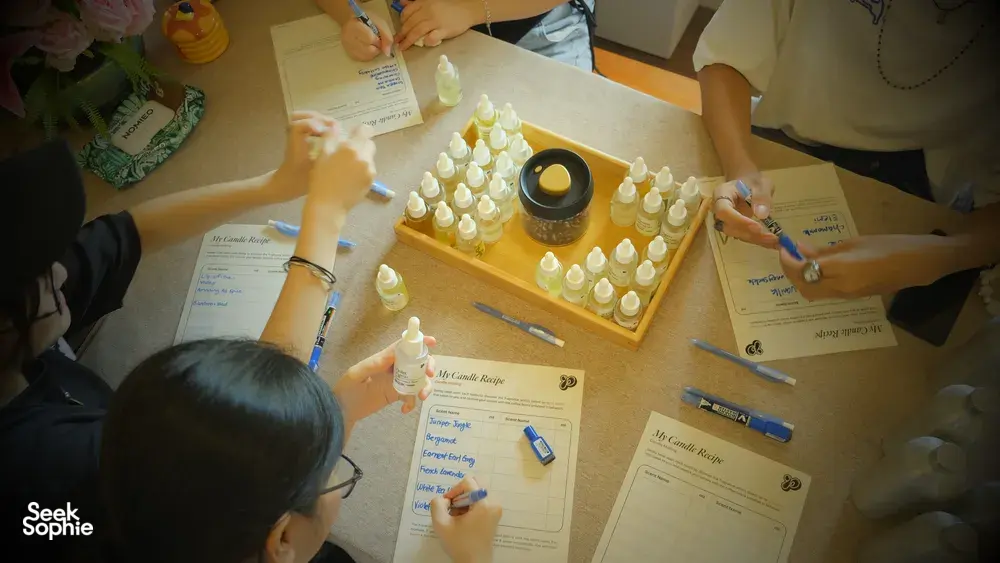Overhead of a Singapore candle-making table with coco-soy jars and droppers, plus scent cards and bottles in a perfume workshop