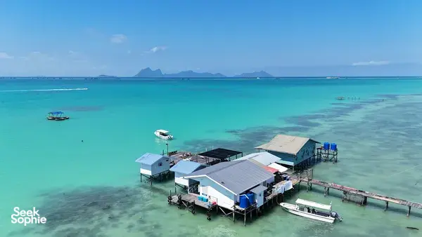 Pemandangan dari udara banglo atas air dan jeti selam di Semporna di atas laut biru firus yang jernih, dengan bot berlabuh dan bukit-bukau di kejauhan