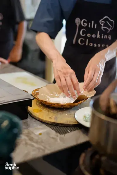 Hands pressing dough in a woven tray at a Gili T cooking class in Lombok, with Gili Cooking Classes aprons