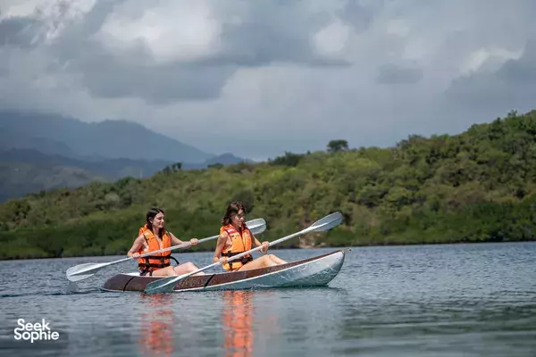 Two paddlers in orange life jackets kayak on a calm Indonesian lake, with green hills and mountains under overcast skies