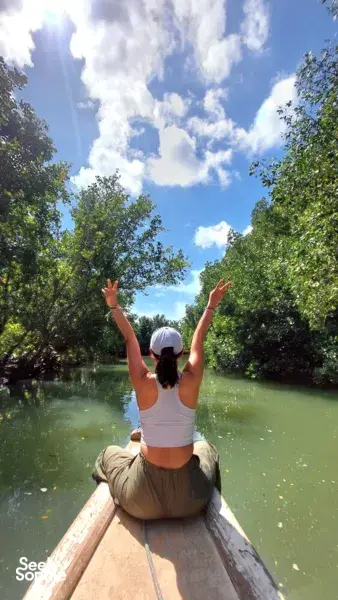 Kneeling in a canoe on calm water through mangroves in Labuan Bajo, arms raised under a bright blue sky