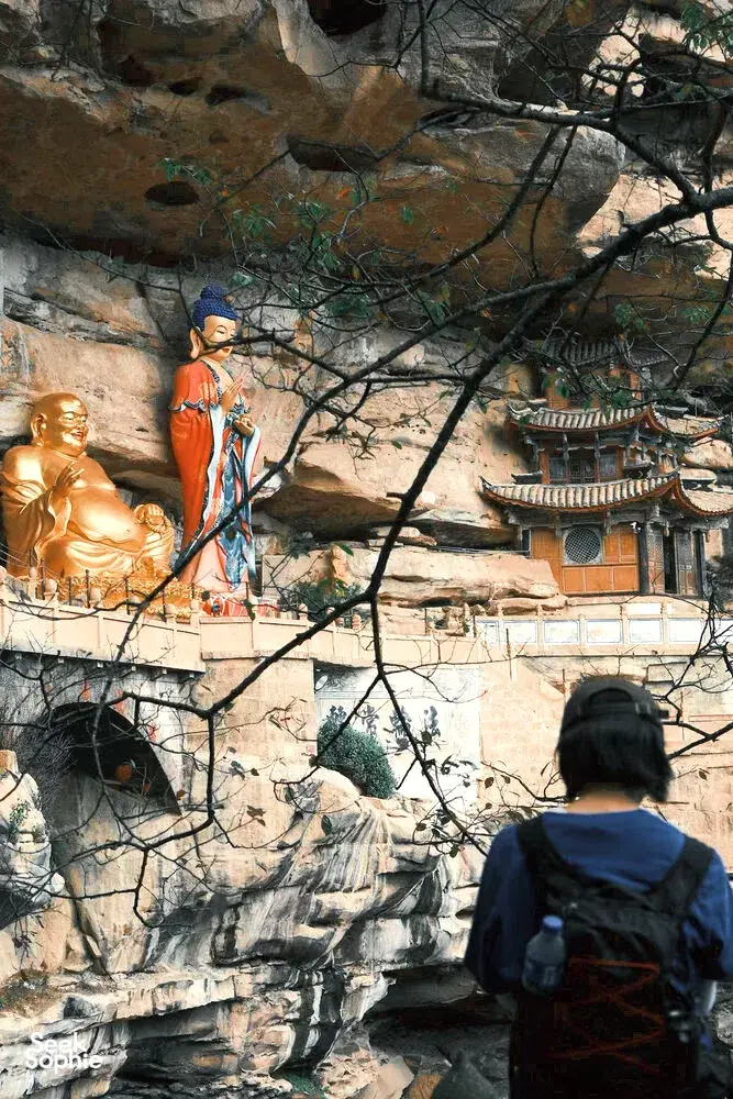 Patung Buddha berwarna dan ukiran gua di gua Buddha, dengan seorang pengunjung di latar depan, eco retreat Shaxi di Dali