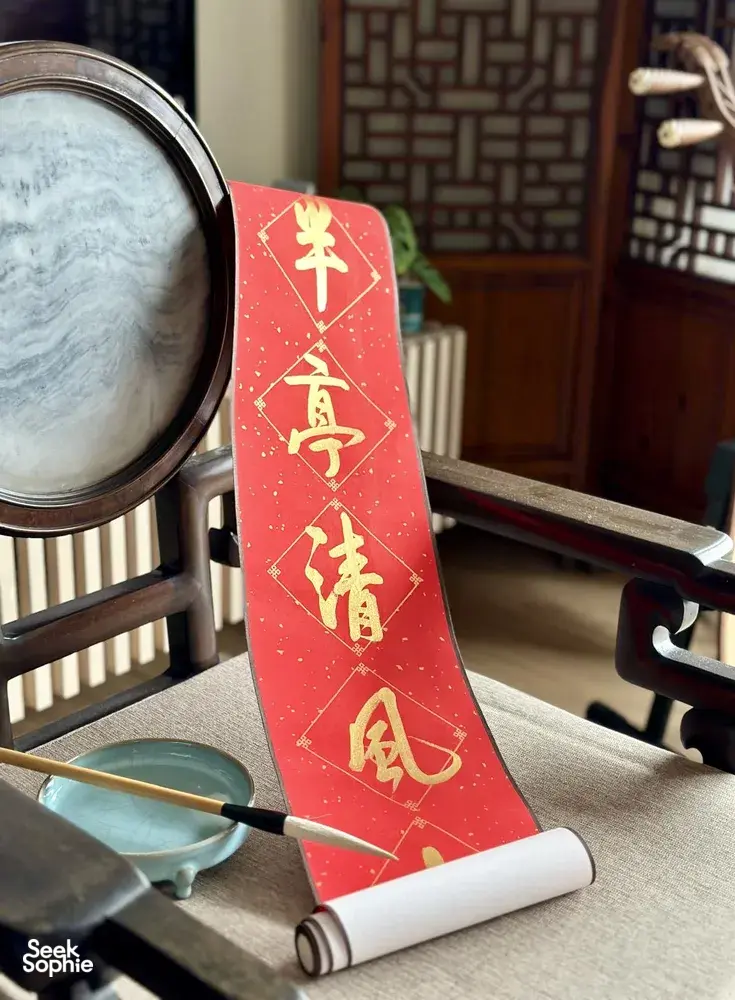 Red Chinese calligraphy strip on a desk with a brush, magnifying glass and rolled practice paper during calligraphy workshops in Singapore