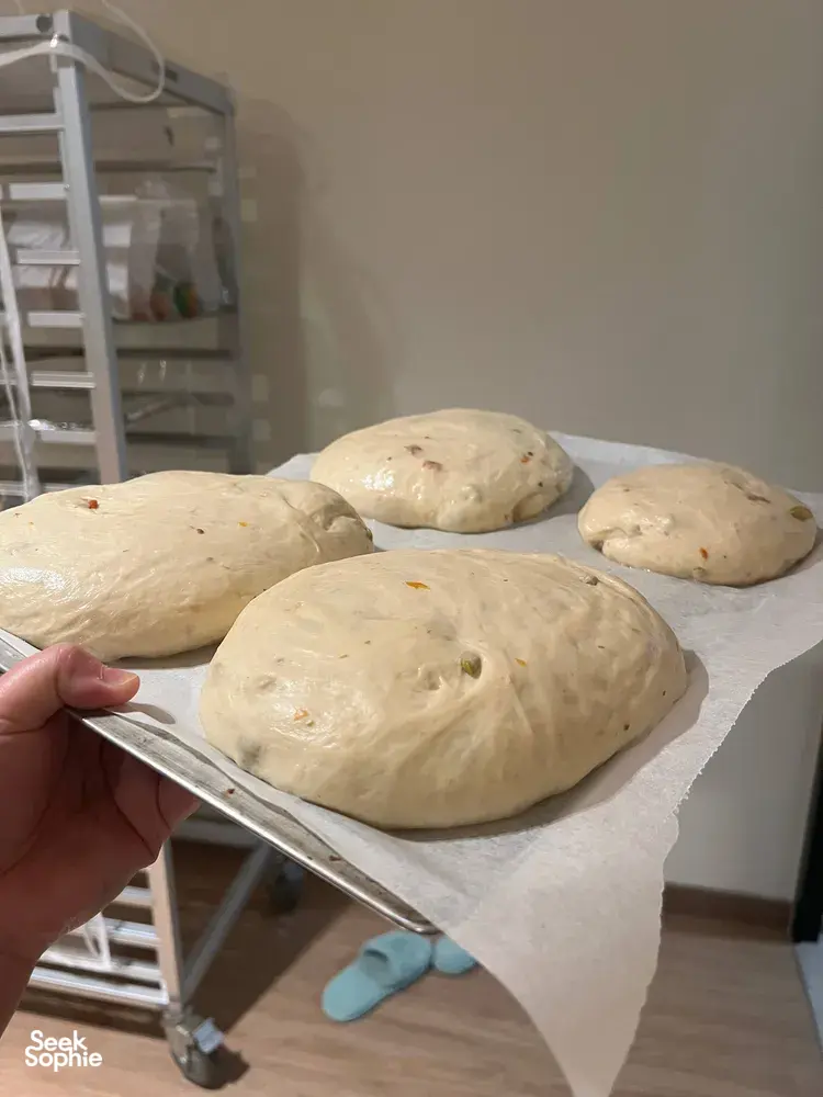 Hand lifting a tray of pale dough loaves waiting to bake in a Singapore kitchen for focaccia making — workshops in Singapore