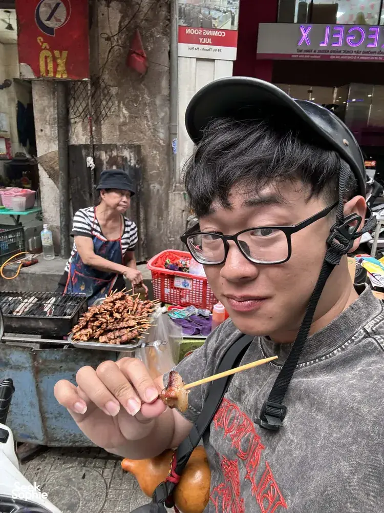 Tour participant wearing a headset eating a grilled skewer at a busy Hanoi street stall, with the grill and ingredients behind
