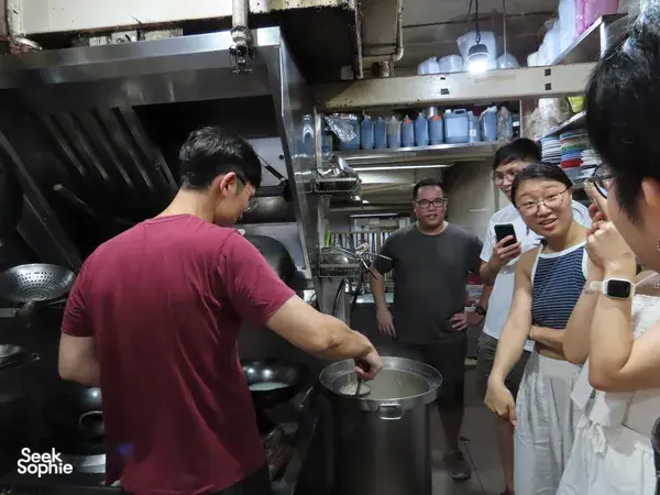 Chef cooking in a Singapore hawker kitchen while a small group watches, part of a Cantonese fish tour and hawker experience