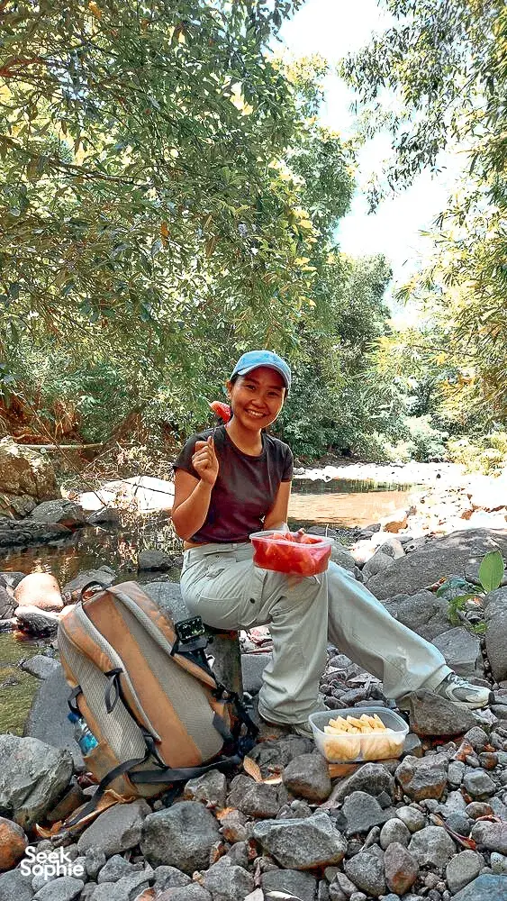 Woman preparing a meal by a rocky stream in the Lombok forest, with bowls and ingredients for the cooking stop