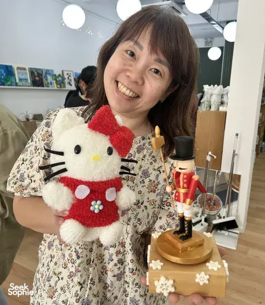 Jan from Sage Artelier smiles while holding a decorated wooden musical-box keepsake beside a nutcracker display, workshops in Singapore