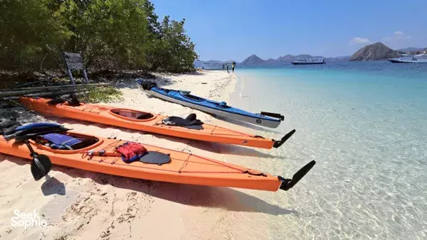 Orange and blue kayaks resting on a sandy shore at Labuan Bajo, with small boats in the calm turquoise bay before paddling
