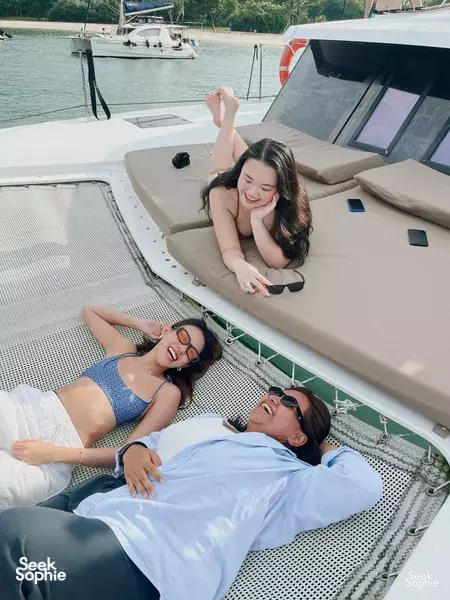 Sunbathers relaxing on cushioned deck seating of a luxury catamaran, with green shoreline and calm sea off Singapore