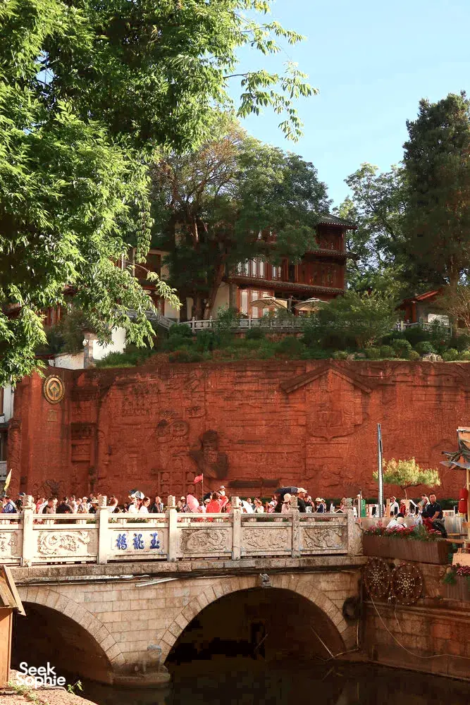 Crowds gathered on a stone bridge over water, with a carved red wall and leafy trees in Lijiang Old Town