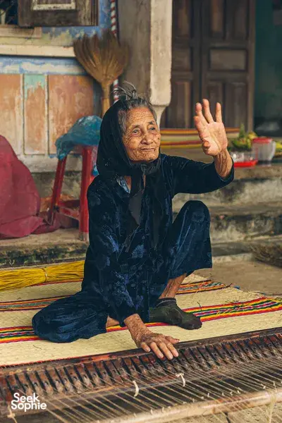 Local artisan seated and waving while weaving on a patterned floor mat in Hoi An, showcasing an old-town craft at dawn