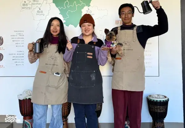 Guests in aprons posing with a small dog in front of a Yunnan coffee map at a coffee workshop in Lijiang