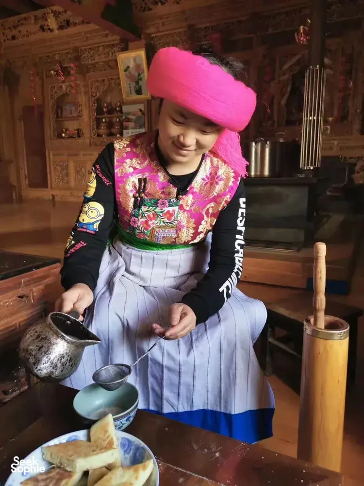 Woman in a pink headscarf preparing food at a hearth in a Tibetan home kitchen in Shangri-La during a host-led lunch