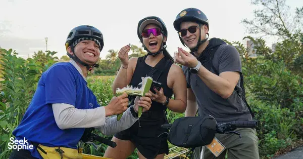 Jackfruit Adventure cyclists in helmets laughing and holding drinks on a bike ride through green paths near Ho Chi Minh City