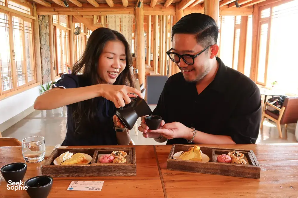 Two people seated at a Lijiang coffee tasting table, pouring coffee from a carafe in a bright wooden workshop