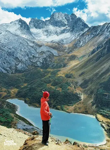 Hiker in a red jacket standing on a steep viewpoint in Shangri-La, looking down at a turquoise river and icy peaks